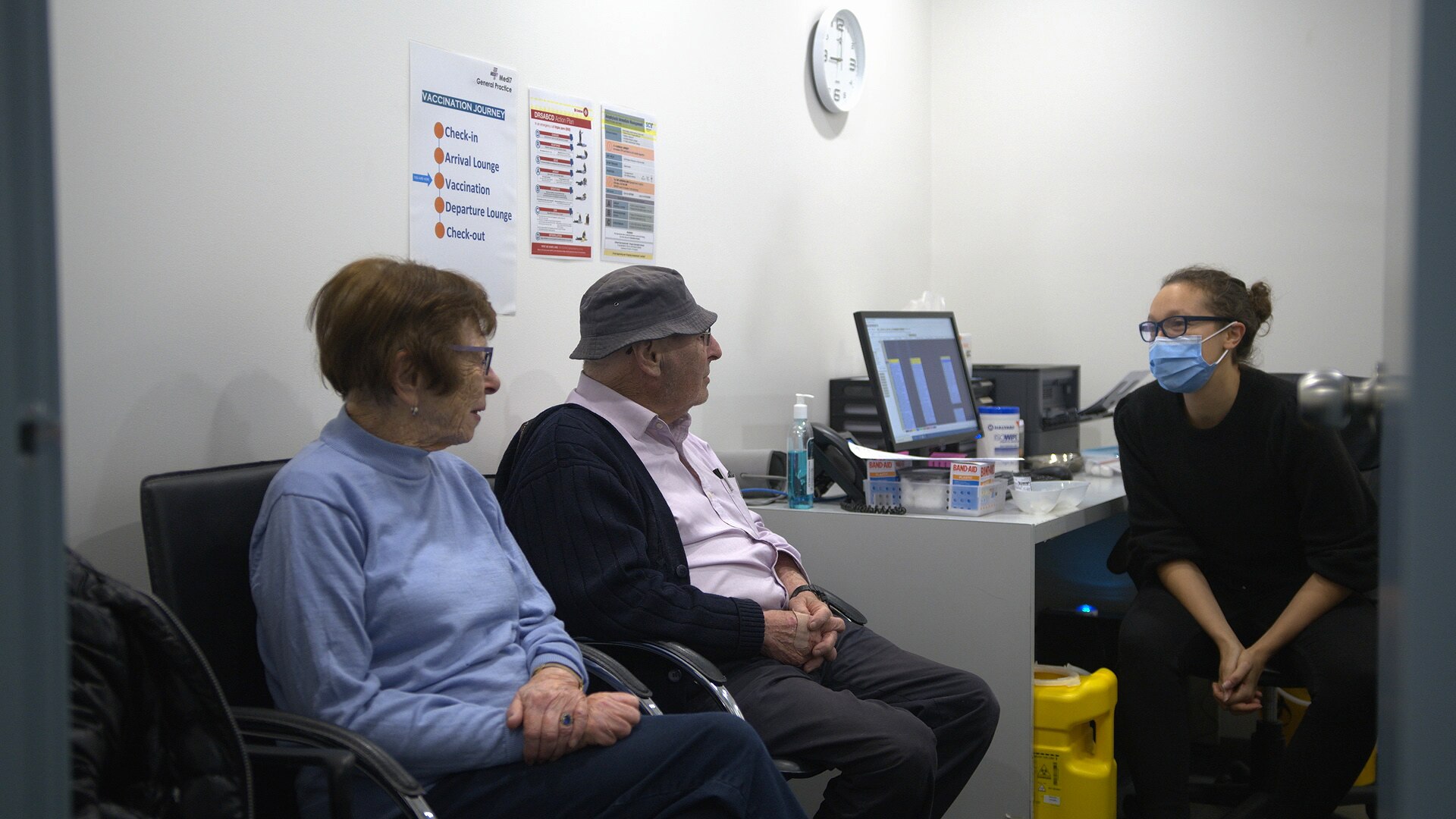 An older man and woman sit in a GP's office talking to a masked doctor.