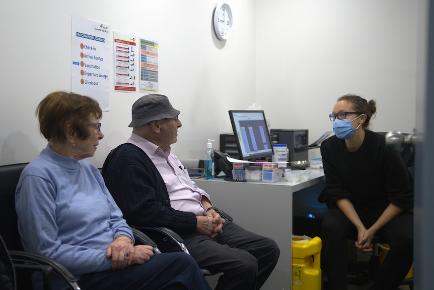 An older man and woman sit in a GP's office talking to a masked doctor.