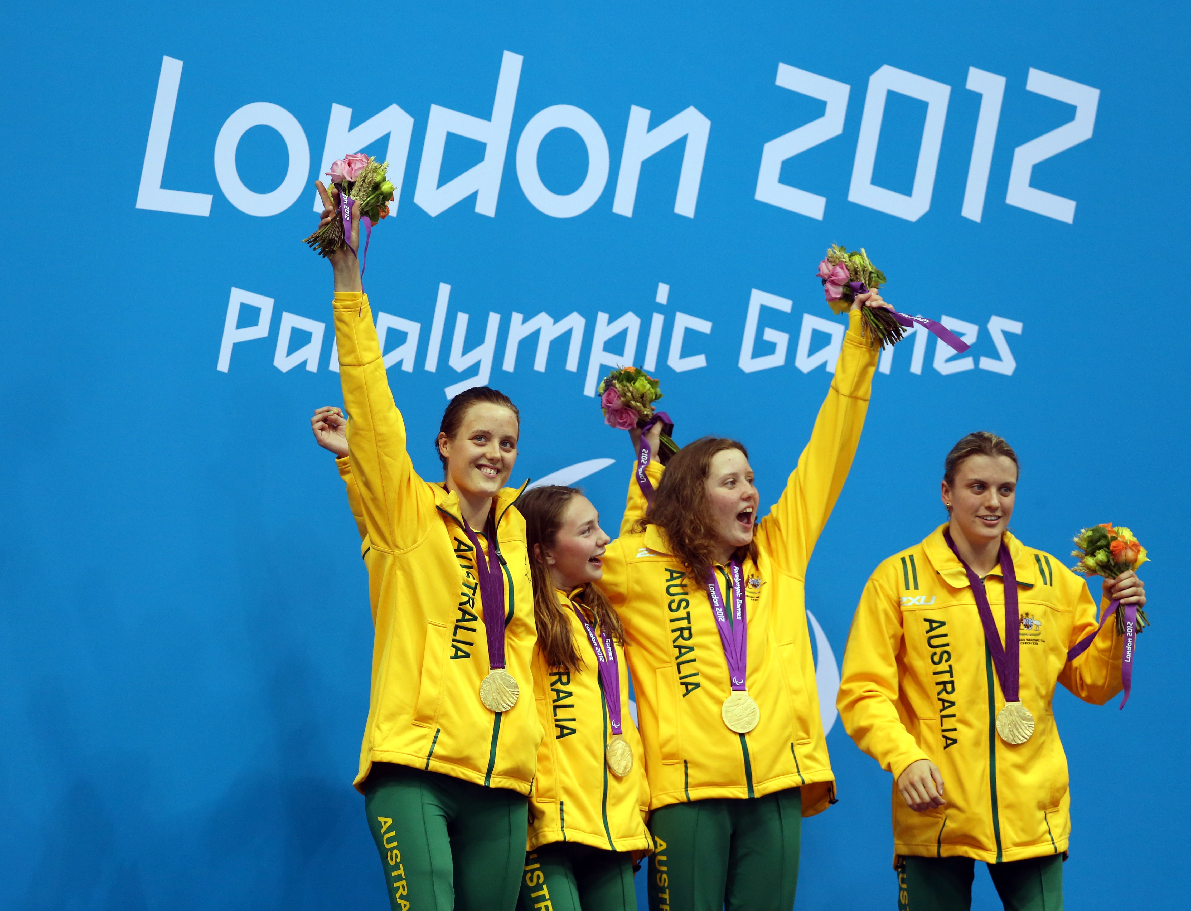 Cuatro mujeres sonrientes que visten el equipo del equipo de Australia con medallas de oro alrededor del cuello sostienen flores mientras están de pie en un podio.