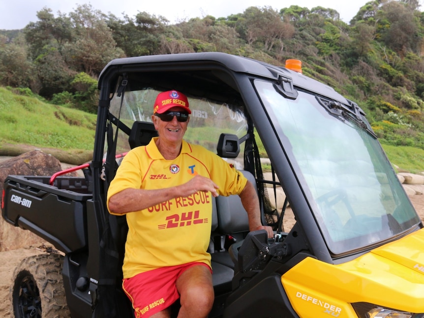 An elderly man wears a surf rescue hat and t-shirt while sitting in a beach buggy.