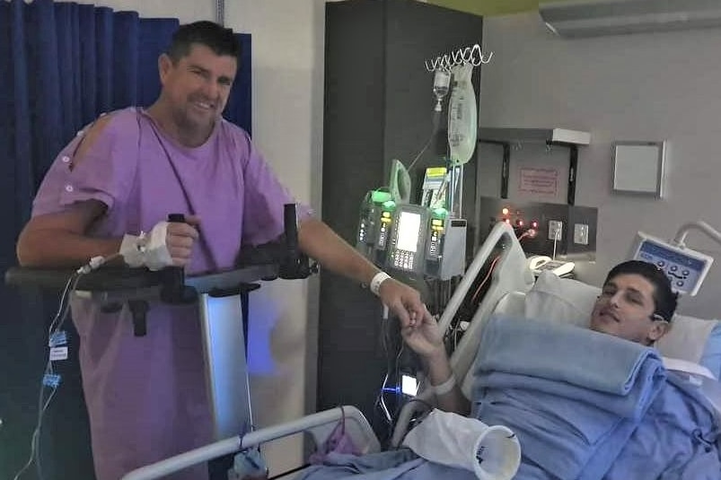 Father and Son holding hands in hopsital. Shane is standing while Bryce is lying in bed. 