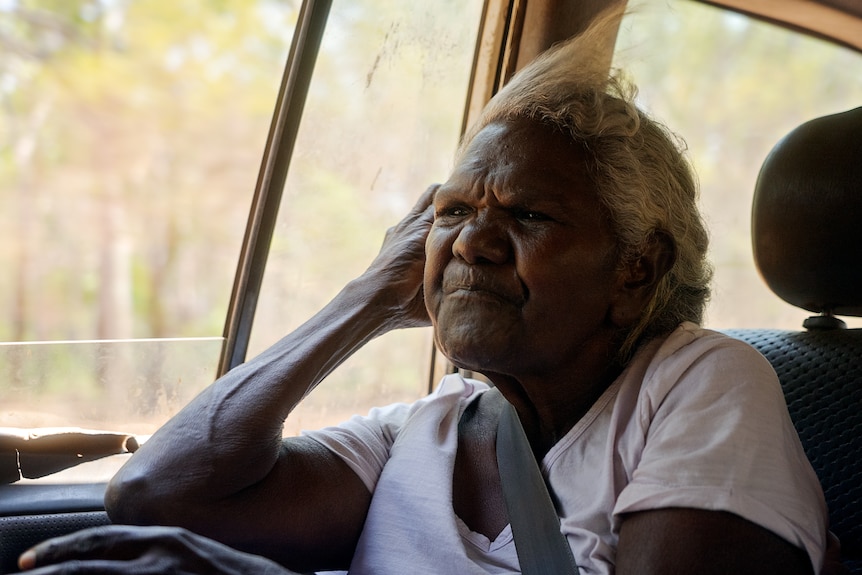 An elderly Indigenous woman sits in a four wheel drive with the window down and wind blowing her greying hair.