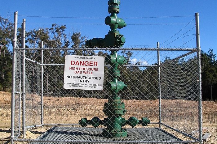 Large green wellhead protruding from the ground, surrounded by a chain-link fence topped with barbed wire