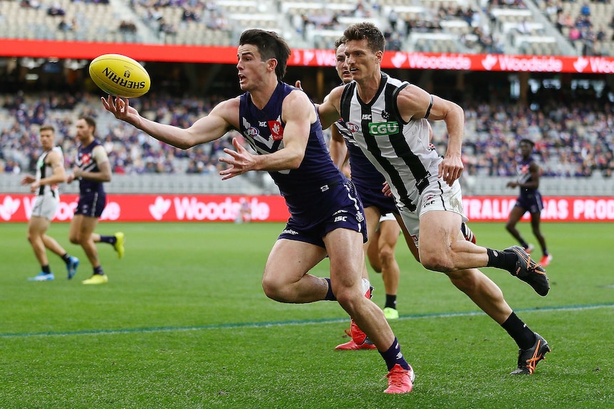 A Fremantle Dockers AFL player attempts to control the ball with his right hand as he is chased by a Collingwood opponent.