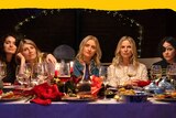 Five white women are seen sitting at a dinner table at night, all except one not smiling.