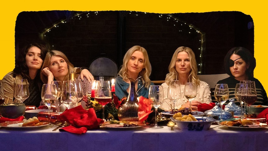 Five white women are seen sitting at a dinner table at night, all except one not smiling.