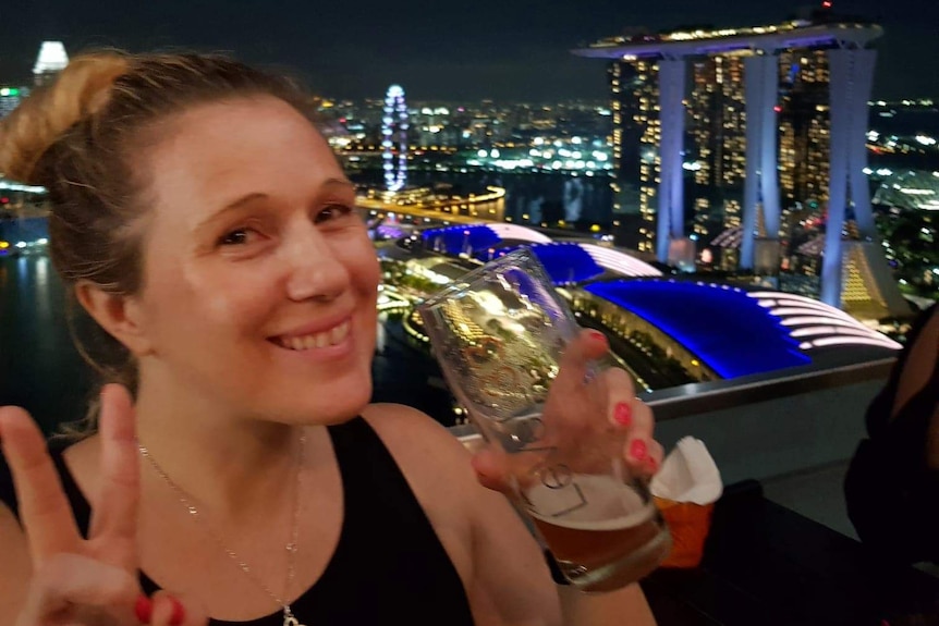 A young woman holds up a peace sign while holding a drink with the Singapore skyline at night behind her.