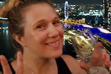 A young woman holds up a peace sign while holding a drink with the Singapore skyline at night behind her.