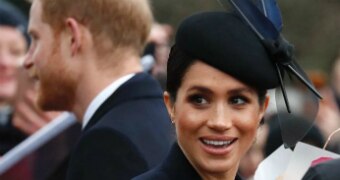 Meghan Markle wears a navy blue fascinator and looks to the camera with a smile on her face with Prince Harry behind her.