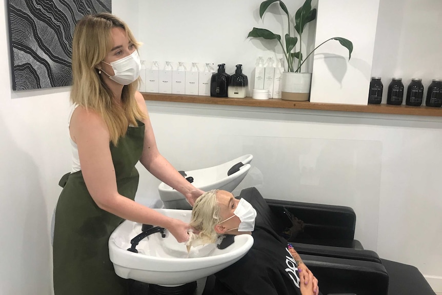 a woman washing the hair of another woman, both are wearing masks