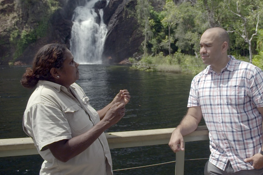 A woman wearing a khaki shirt talks and gestures to a man wearing a checked shirt, with a waterfall behind them.