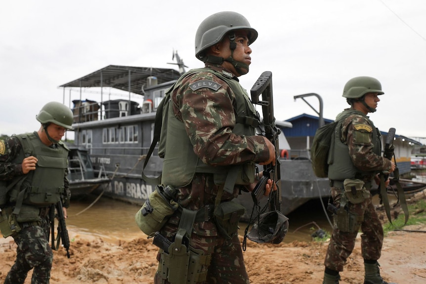 三名身穿制服的士兵在沙滩上携带武器，背景是一条深棕色的河流