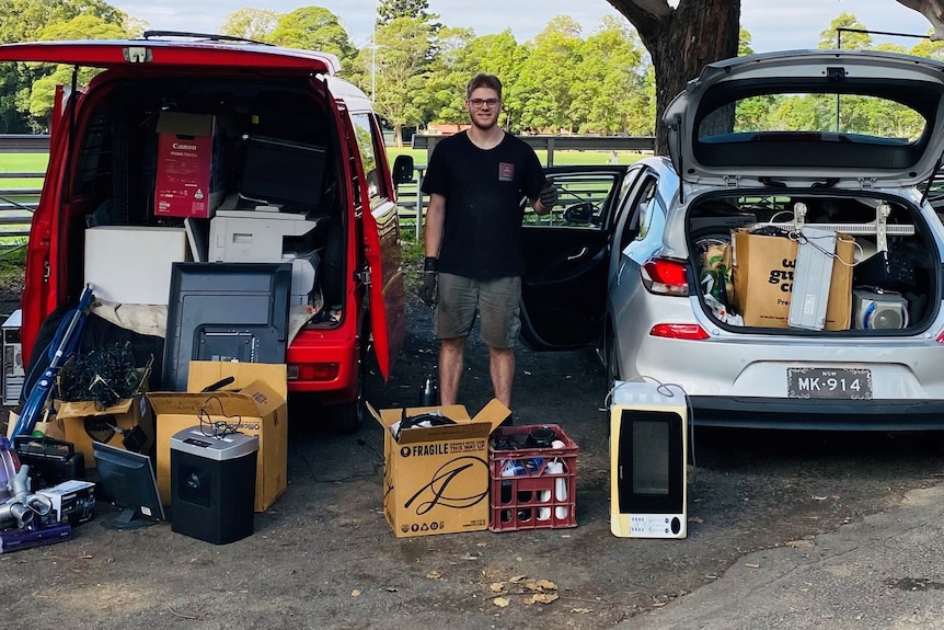 Wade wears a black t-shirt and grey shorts, and is standing between a red van and a silver car full of old electronics. 