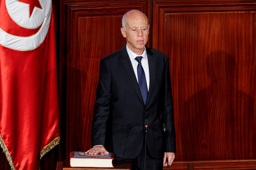 A man in a suit stands next to the red Tunisian flag with his hand on a book swearing an oath