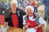 Two smiling women, both wearing red aprons with 702 Sydney ABC on it, one has grey hair, mixes ingredients in a bowl.