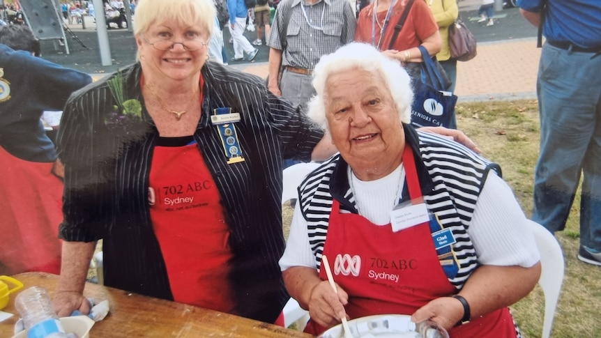 Two smiling women, both wearing red aprons with 702 Sydney ABC on it, one has grey hair, mixes ingredients in a bowl.