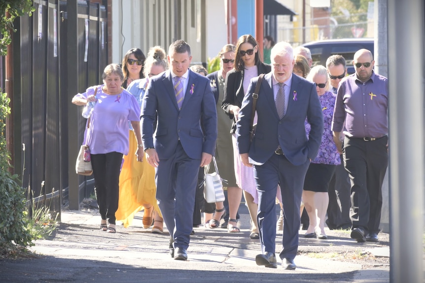 Two men in suits walking with a pack of people in purple and yellow dress