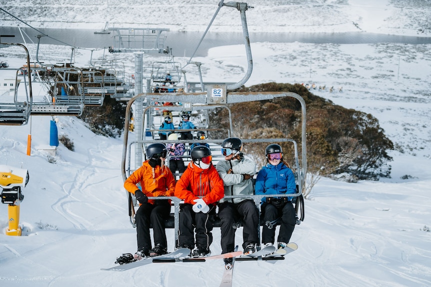 A group of four people riding a ski chair lift with a snowy backdrop and other occupied chair lifts in the background