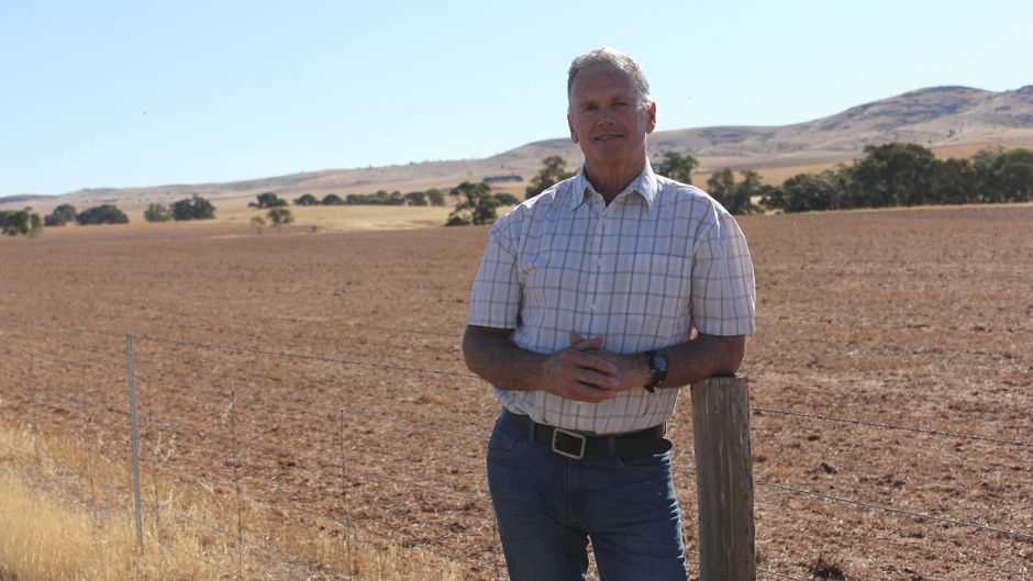 A man in button-up shirt and jeans leaning on a fence post with the Flinders Ranges in the background.