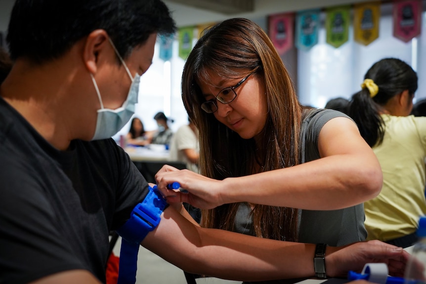 A woman wearing grey shirt applies a torniquet to the upper arm of a man in face mask