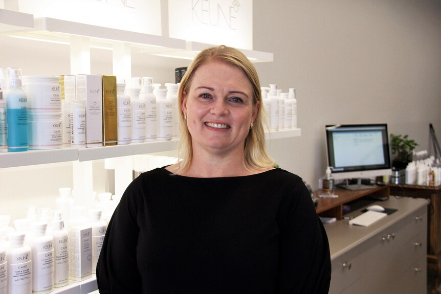 A blond woman in a black top smiles in front of shelves of hair care.