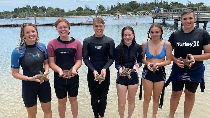 A group of high school girls and boys stand in wetsuits on the shore of a lake, holding starfish.