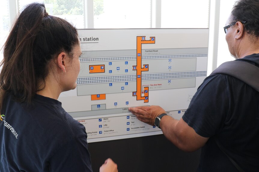 two people standing by a braille map of a train station. one woman is using her fingers to read it