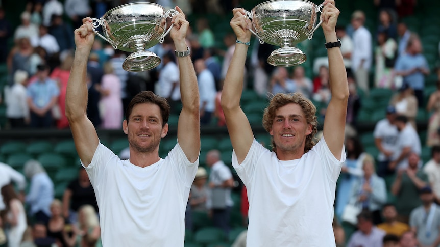 Two men in tennis whites smile and hold trophies aloft