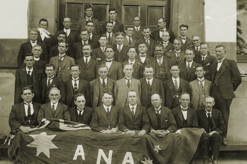 An old black and white photograph of a group of men in suits with a flag saying ANA