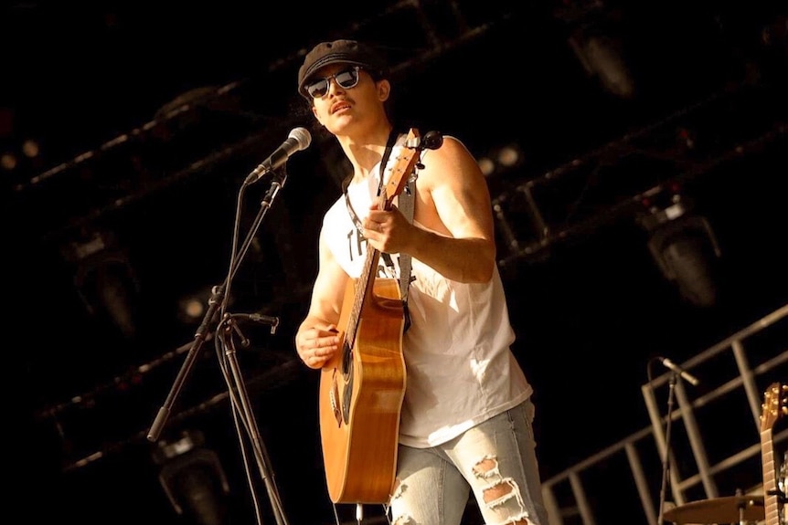 A young man in a cap and sunglasses plays an acoustic guitar onstage.