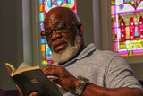 Jerry Givens sits in a chair in front of stain-glass windows reading a Bible.