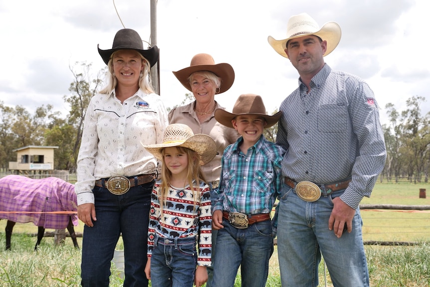 Three adults and two kids stand in front of a horse paddock wearing big hats smiling at the camera