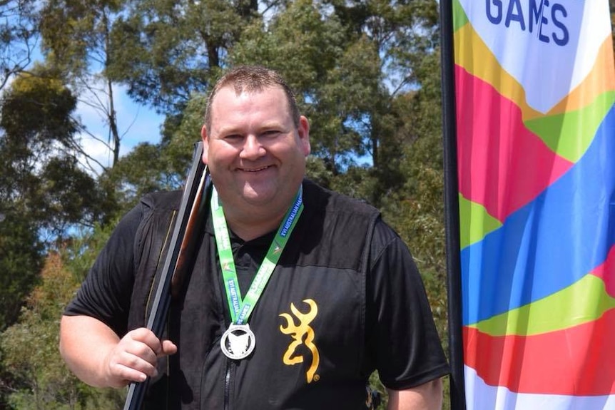Man standing holding a gun over his shoulder, wearing a medal and standing next to a sign advertising the Masters Games