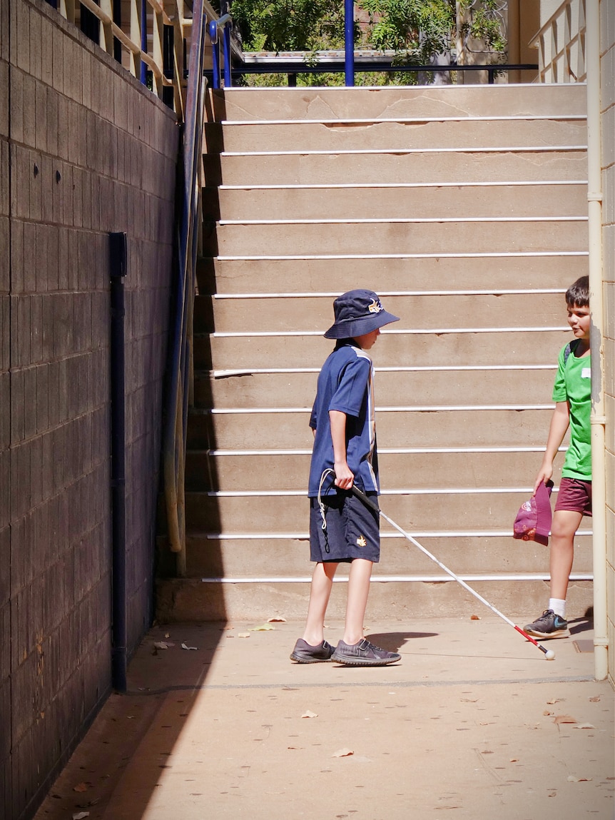 Young boy carries white cane walking on school pavement at bottom of stairs