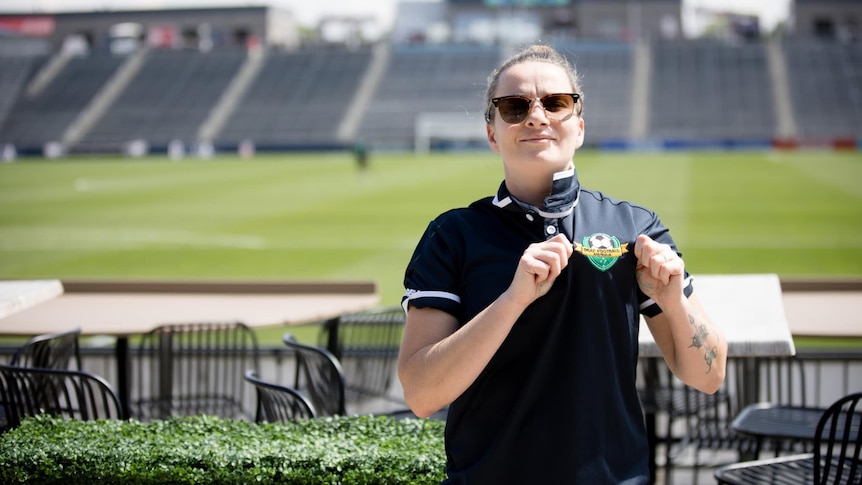 A white woman wearing sunglasses holds her polo shirt which has a soccer ball logo, she's standing in front of a soccer pitch