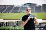 A white woman wearing sunglasses holds her polo shirt which has a soccer ball logo, she's standing in front of a soccer pitch