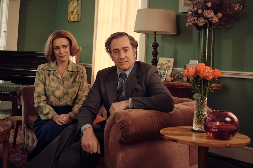 A male and female actor pose for a photo, dressed smartly, on a sofa in a 70s style living room with green walls