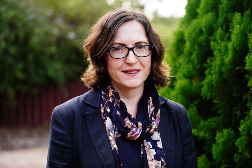 Kirstin Holmes smiling in a courtyard with green plants in soft focus in the background.