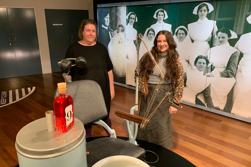 Two gallery staff members standing behind a blue and grey antique dentist chair with giant historic photo of nurses on the wall
