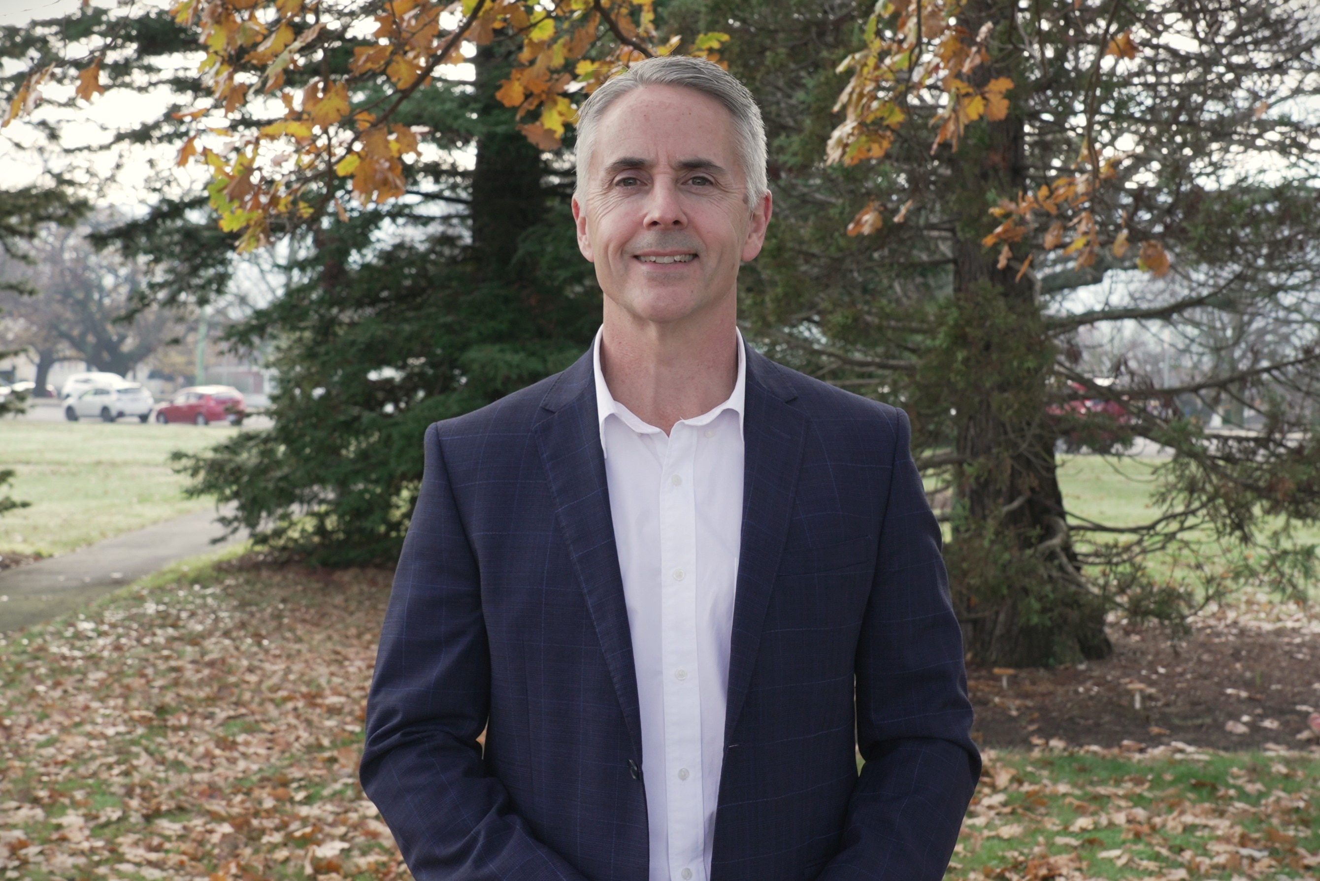 Nick Steel stands in a public park wearing a blue and white suit, smiling into the camera.