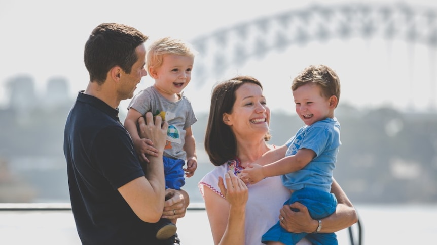 Two parents hold two young boys in front of the Harbour Bridge