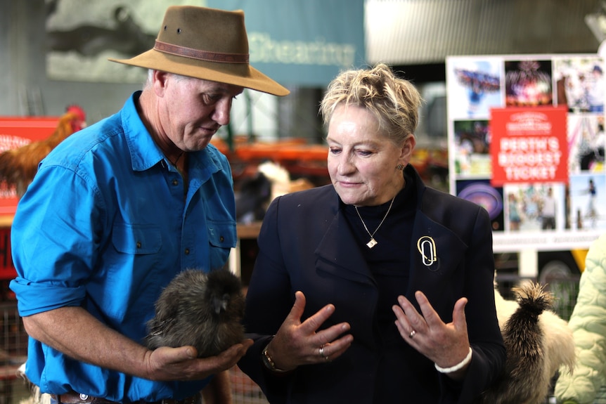 A well-dressed woman looks at a bid being held by a man dressed in denim and a broad-brimmed hat. 