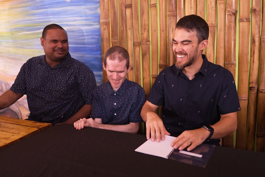 Three men sit at a dining table with a dark table cloth. All are smiling and one man is reading a menu in braille