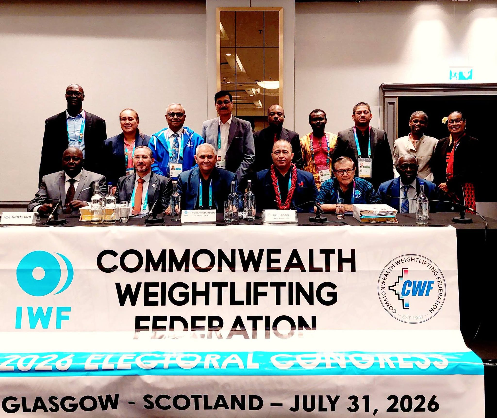 A Samoan man sits at a table with the words Commonwealth Weighlifting Federation on a banner hanging in front of the table