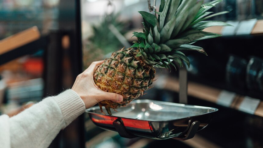 A woman places a pineapple onto a set of scales in a store