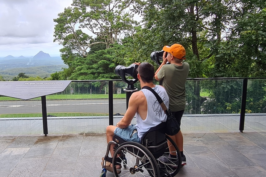 A man in a wheelchair sits looking through lookout binoculars with a boy standing beside him also looking through a binocular