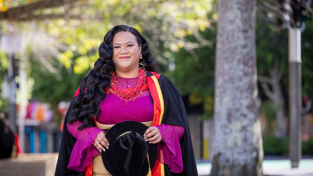 Tongan woman Linda smiles outdoors with luscious long black hair swept to once side in a purple dress and doctoral gown holding 