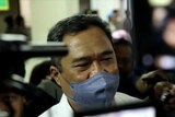 A middle-aged Indonesian man in a white business shirt and a blue mask speaks to a gaggle of reporters in a courthouse.