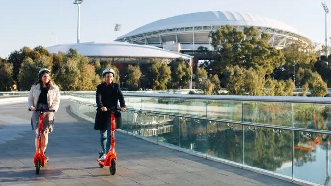a woman and man on orange e-scooters travelling across pedestrian bridge with adelaide oval in background