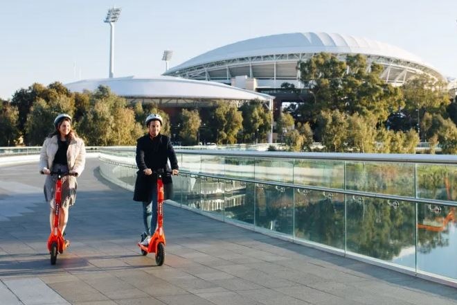 a woman and man on orange e-scooters travelling across pedestrian bridge with adelaide oval in background